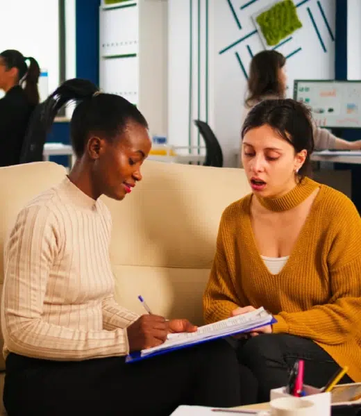 Two women on a couch, one writing on a clipboard, the other talking.